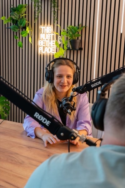 Smiling podcaster with headphones at the microphone, neon sign and plants behind her